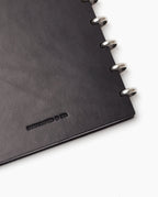 Close-up of the corner of a black Discbound Heirloom Journal (Standard 6" x 8.5") with full-grain leather and silver metal rings; HANDCRAFTED IN USA is embossed on the cover.