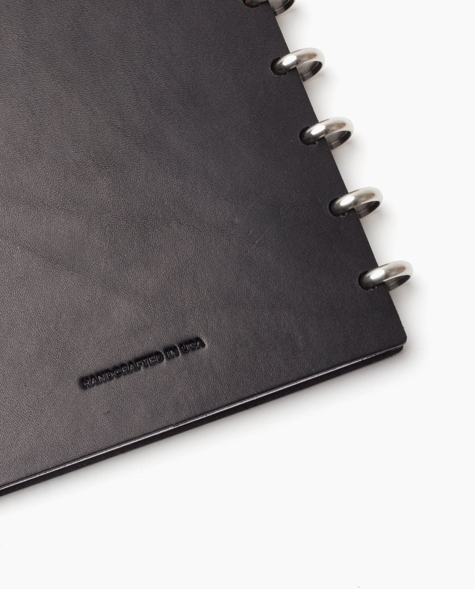 Close-up of the corner of a black Discbound Heirloom Journal (Standard 6" x 8.5") with full-grain leather and silver metal rings; HANDCRAFTED IN USA is embossed on the cover.