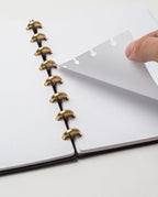 A hand lifts a dot-grid page from the Discbound Heirloom Journal Bundle (Black Leather - Standard 6" x 8.5" + 3 Refills), open on a white surface with gold disc binding and removable, repositionable pages.