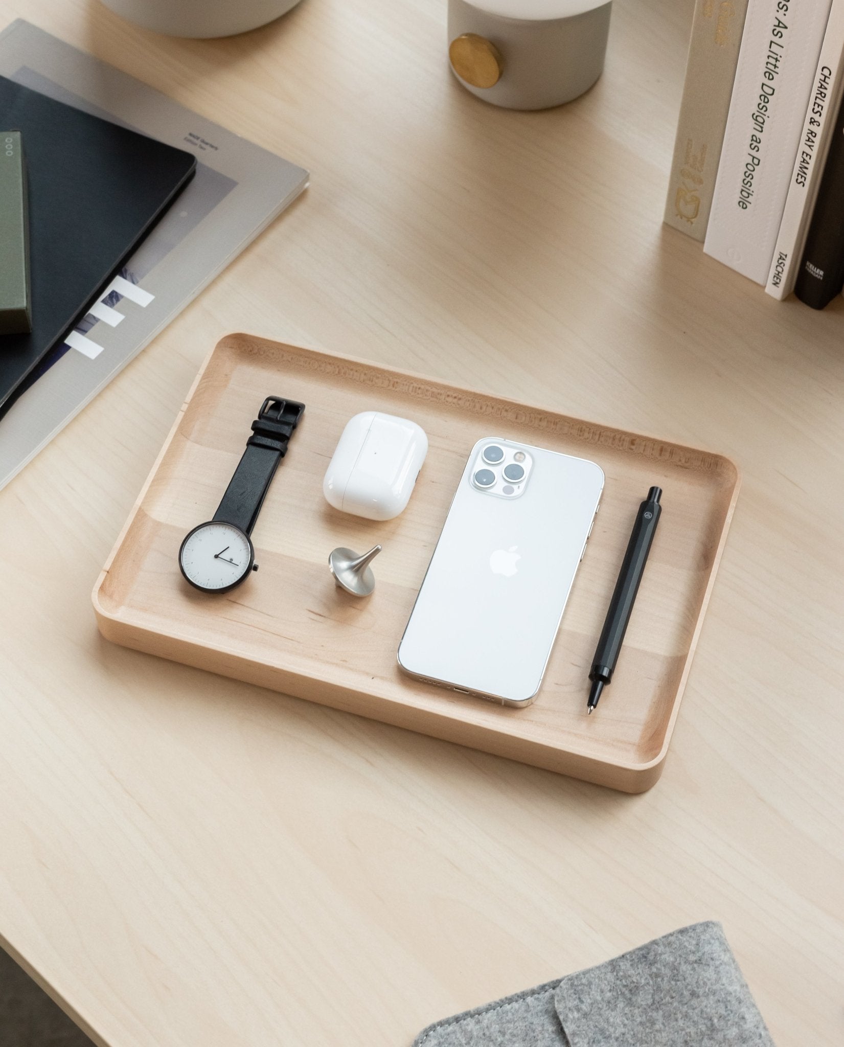 A handcrafted Valet Tray Bundle (Maple - Set of 3) from Pennsylvania sits on a desk holding a black wristwatch, wireless earbuds case, spinning top, white smartphone, and black pen. Books and desk items can be seen in the background.