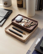 Two handcrafted Valet Tray (Maple - Medium) trays on a desk neatly hold pens, keys, coins, a keychain, and wireless earbuds. Nearby are a felt pouch, white bottle, and books, creating an organized workspace atmosphere.