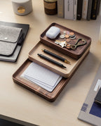 The Valet Tray (Walnut - Small), handcrafted in Pennsylvania, neatly holds coins, keys, a keychain, earbuds, two pens, and note cards on a tidy desk alongside books, a notebook, and a coffee cup.