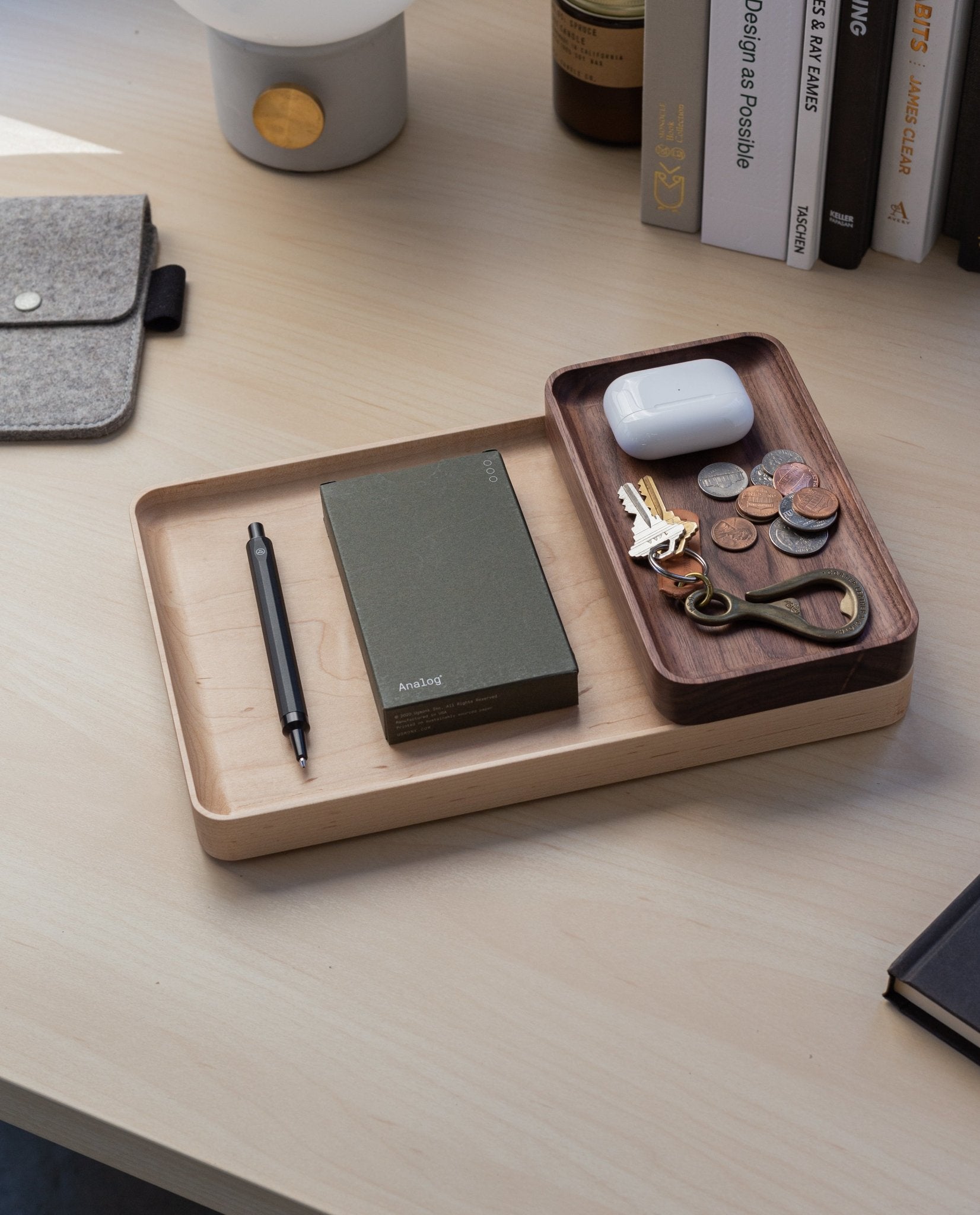 The Valet Tray (Walnut - Small), handcrafted in Pennsylvania, keeps your pen, green notebook, bottle opener, keys, coins, and white wireless earbuds organized on your desk—ideal for everyday carry storage near books and a gray felt pouch.