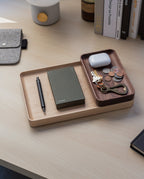 The Valet Tray Bundle (Maple - Set of 3), handcrafted in Pennsylvania, neatly holds a pen, green notebook, keys, coins, a brass bottle opener, and white wireless earbuds case on a light wood desk beside books and a gray fabric pouch.